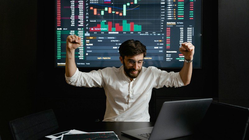 Businessman celebrates stock market success with hands raised in excitement at a trading desk., tags: cs2 - pexel
